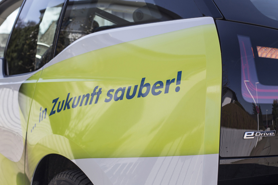 Seitliche Nahaufnahme eines hellgrün beklebten HappyMaids Servicewagens mit Aufschrift ‚Zukunft sauber!‘ und eDrive-Logo