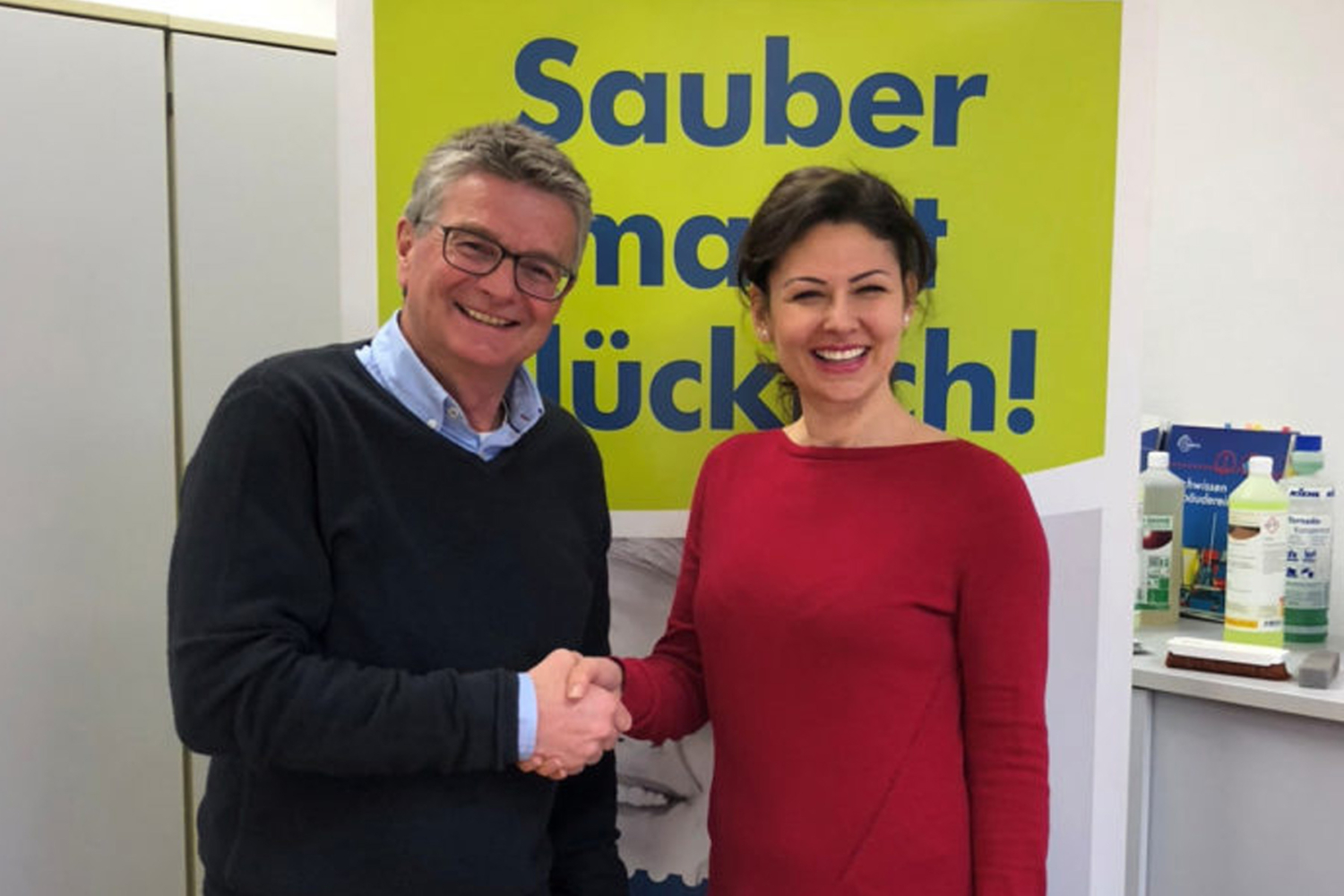 Zwei lächelnde Personen schütteln sich die Hand vor einem HappyMaids-Poster mit Reinigungsprodukten im Hintergrund – Übergabe an neuen Franchisepartner