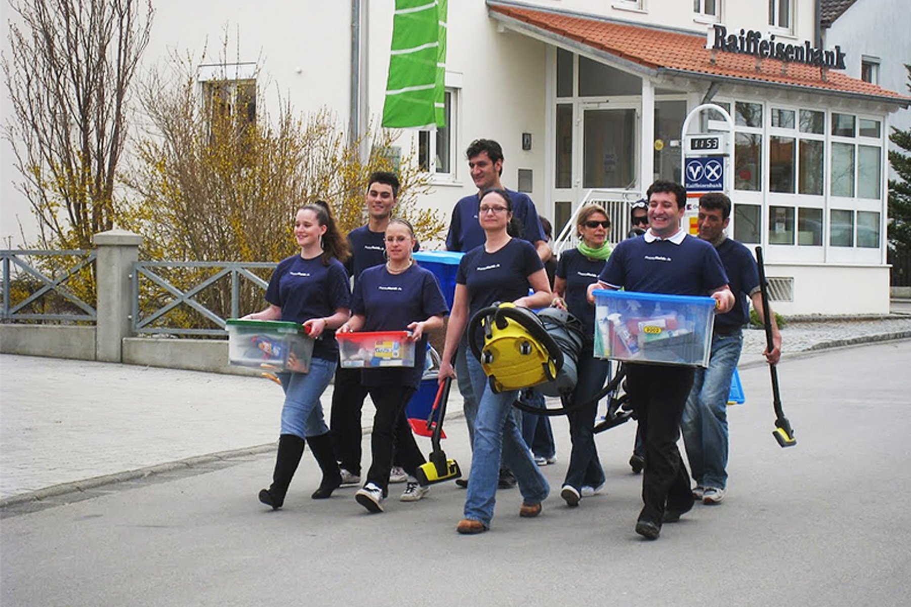 HappyMaids Franchisepartner in einheitlichen T‑Shirts tragen Reinigungsgeräte und Kisten bei einem Partnertreffen auf der Straße vor einem Gebäude