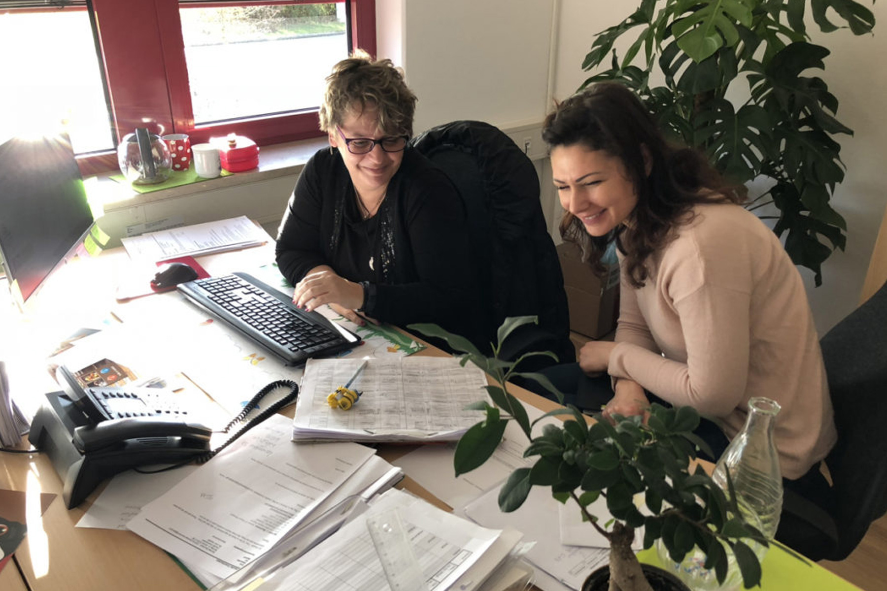 Zwei lächelnde Frauen am Schreibtisch mit Tastatur, Unterlagen und Pflanzen bei einem HappyMaids Franchisepartner-Partnertreffen und Schulung