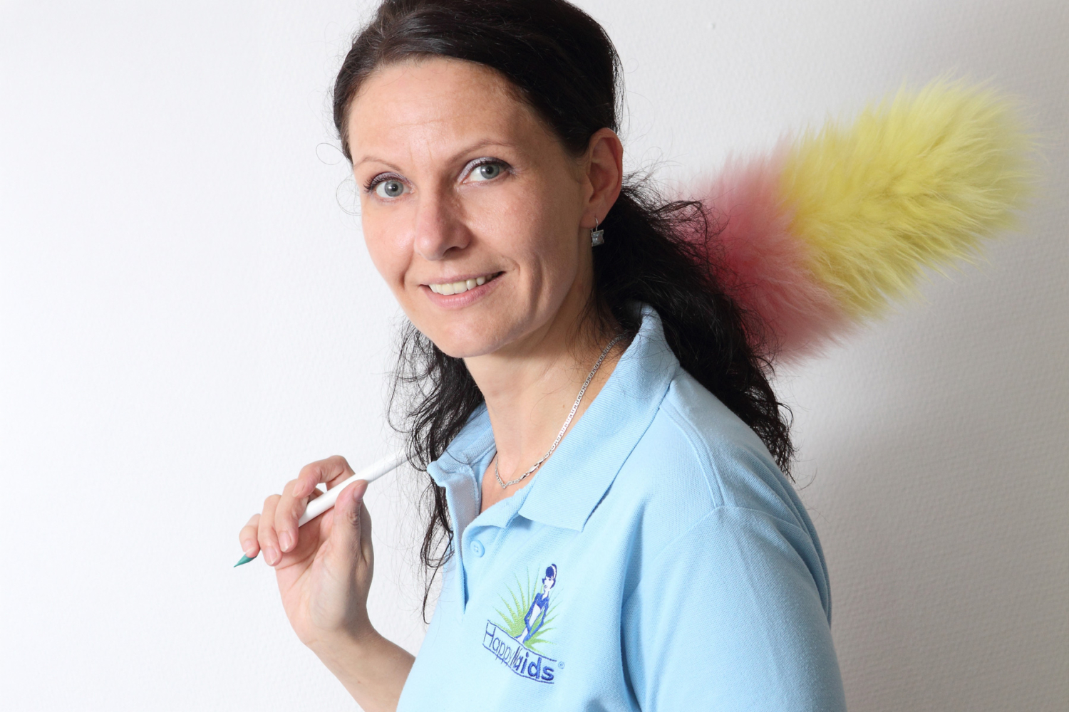 HappyMaids Mitarbeiterin in blauem Poloshirt mit buntem Staubwedel über der Schulter, lächelndes Porträt