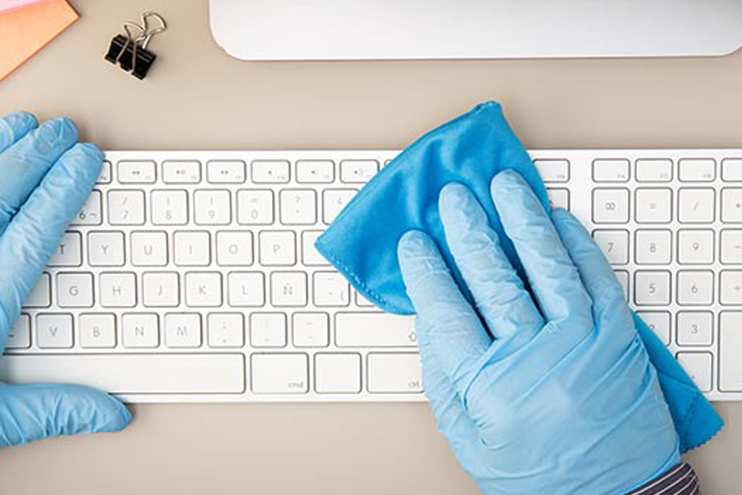 Büroreinigung: Hand mit blauen Einweghandschuhen reinigt weiße Tastatur mit Mikrofasertuch – happymaids professionelle Büroreinigung