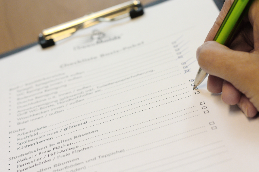 Hand mit Stift füllt Checkliste auf Klemmbrett aus, Reinigungspunkte auf Putzplan für HappyMaids