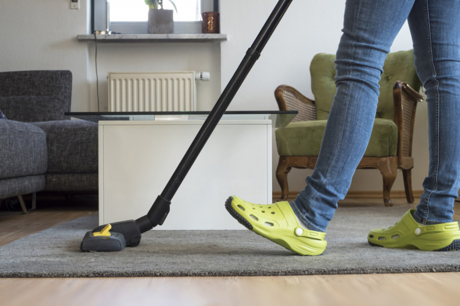 Person mit grünen Hausschuhen beim Staubsaugen im Wohnzimmer, HappyMaids Reinigung mit Staubsauger auf Teppich vor Sofa und Sessel