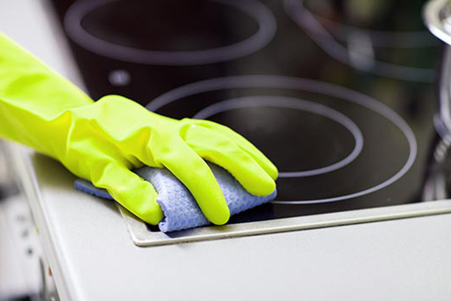 Hand mit gelbem Reinigungshandschuh wischt Cerankochfeld mit Tuch – HappyMaids VIP Paket professionelle Wohnungsreinigung